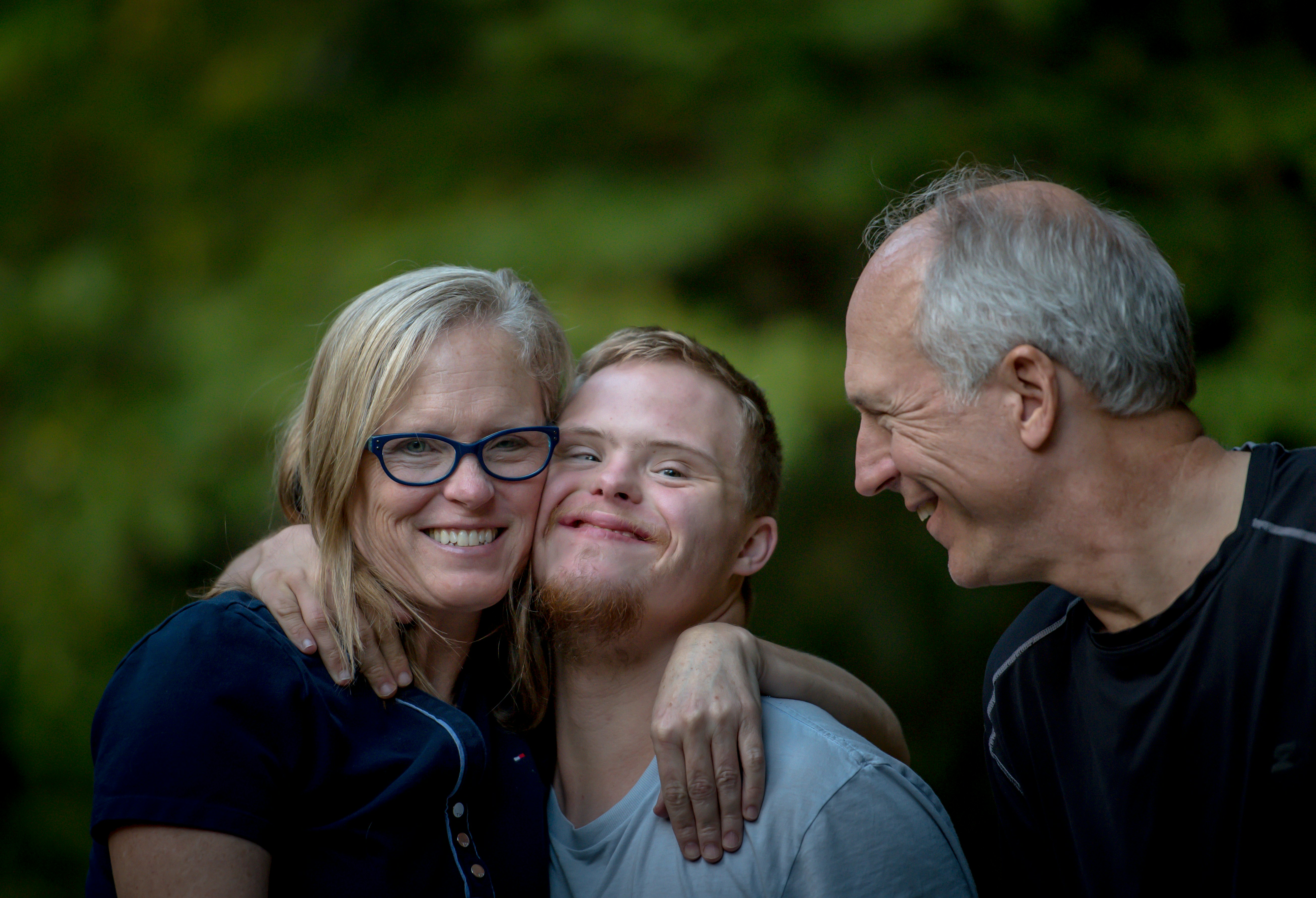 To the left, a woman hugs her son as they smile while her husband to the right of them looks fondly at them.