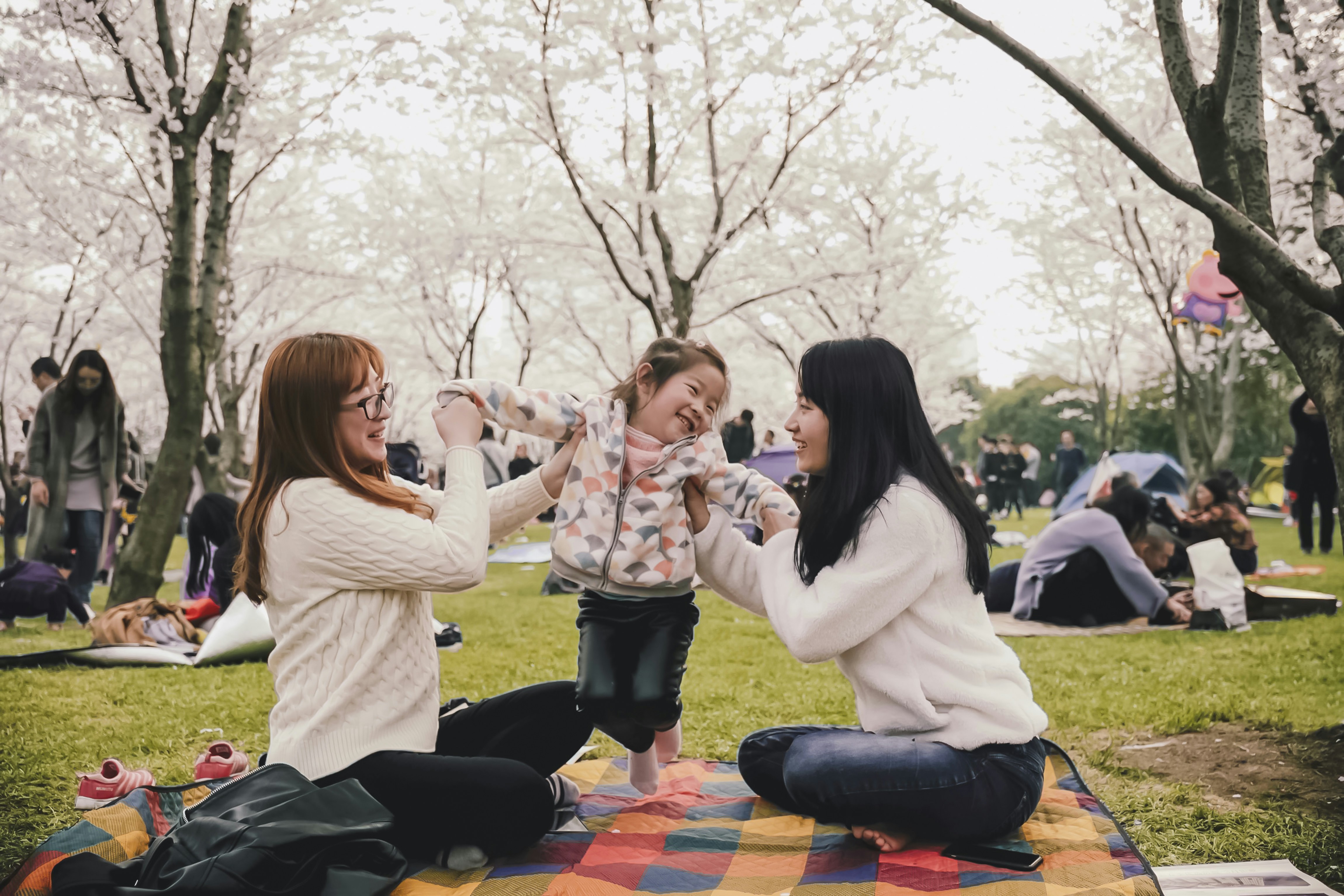 Two women sitting across from each other lift up their daughter between them while relaxing at the park.